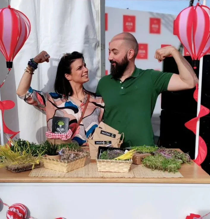 Osnivači brenda MOĆNO (Milena Jovanović i Ivan Jovanović) pokazuju mišiće pored štanda sa mikrobiljem, simbolizujući snagu i zdravlje koje njihovi proizvodi donose.