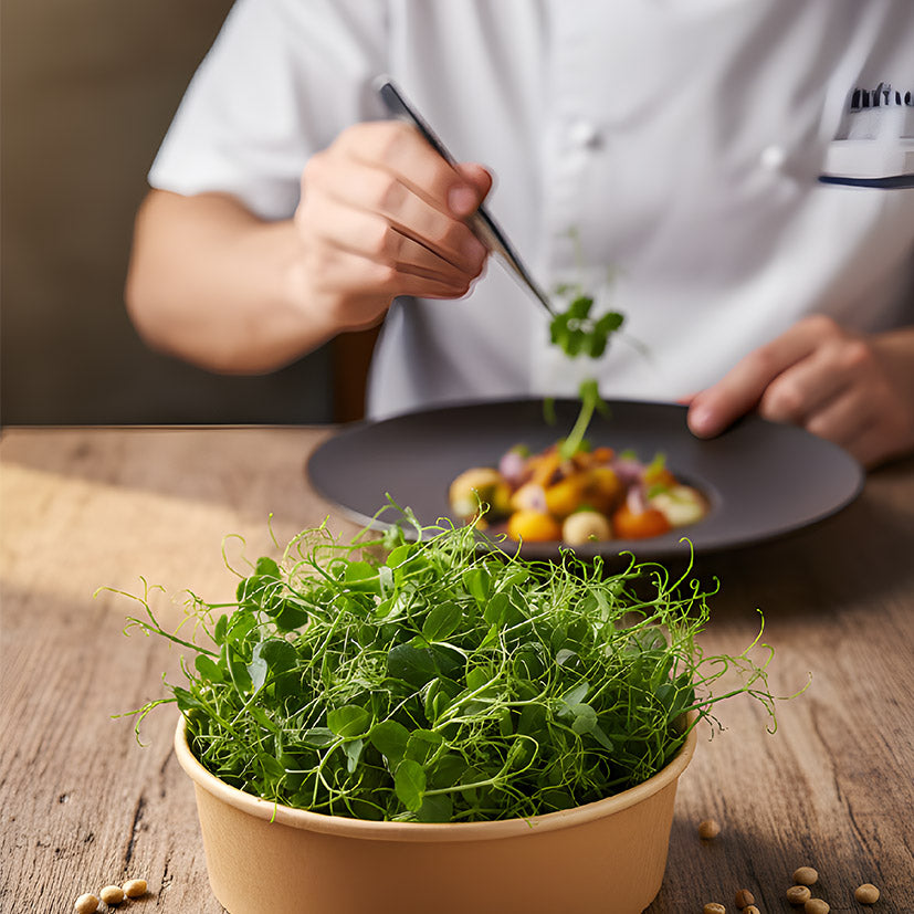 A close-up of fresh MOĆNO pea shoots in a round eco-bowl on a wooden table, with a professional chef using tweezers to plate a dish in the background.